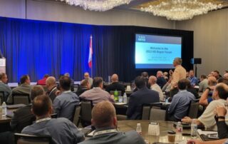 A large audience at the 2022 Repair Forum with a speaker at the podium, featuring flags and a presentation screen.